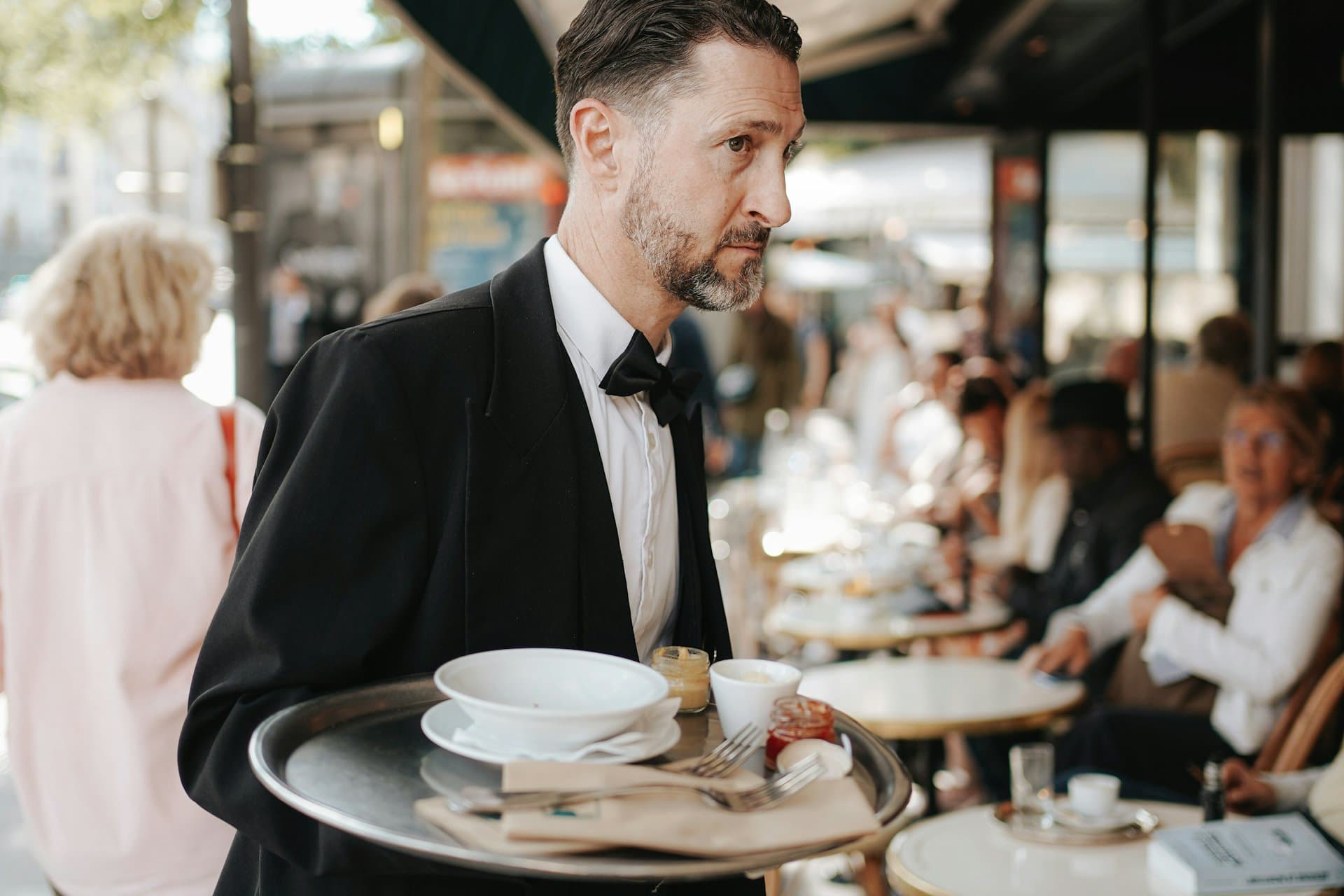 A waiter carrying a loaded tray through a busy pavement café.