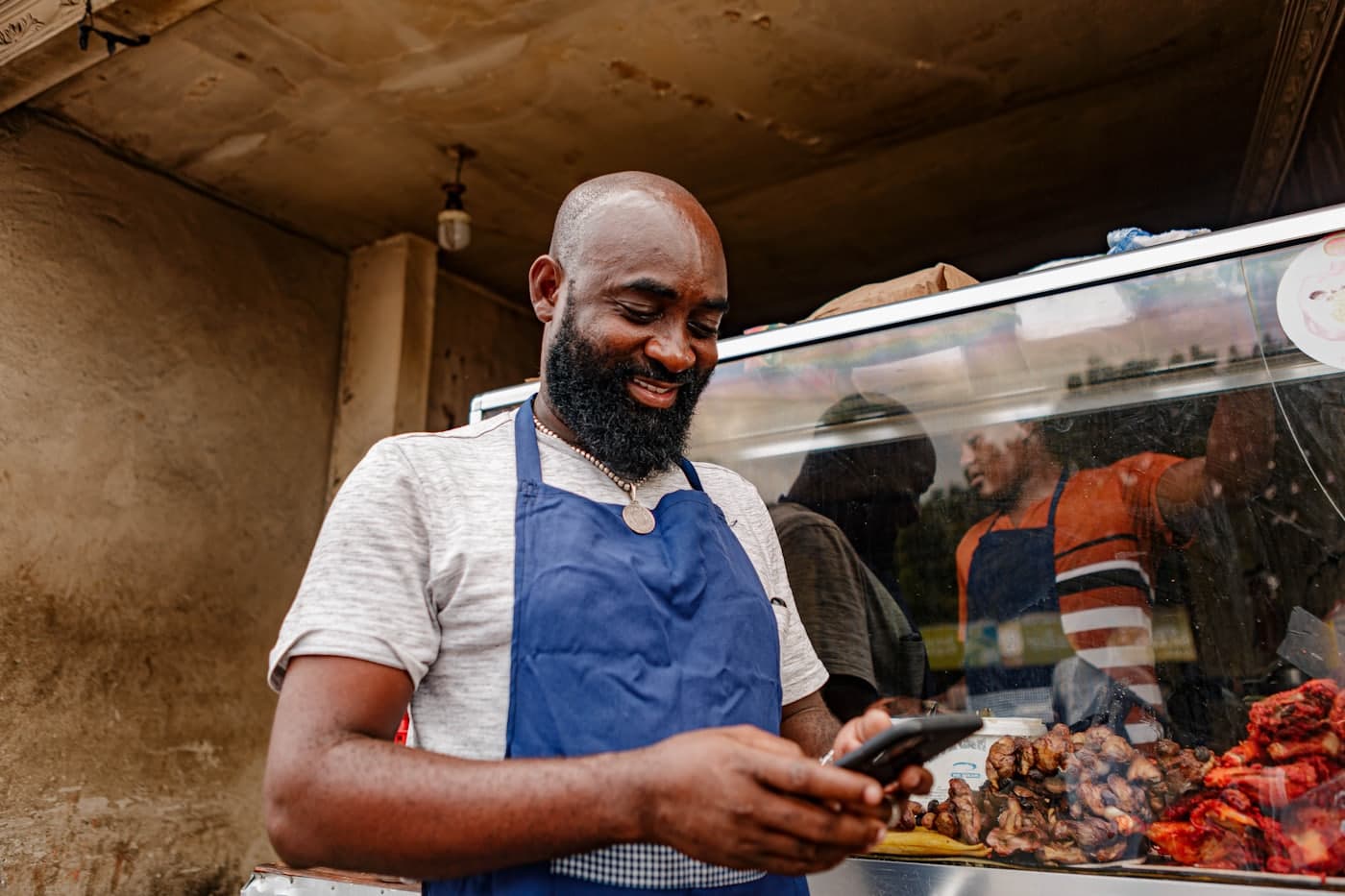 A shop owner in an apron smiling at his phone behind the counter of his food business.